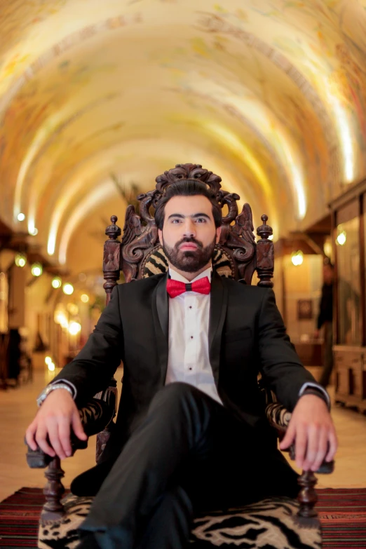 a man in a tuxedo sitting in a chair, an album cover, inspired by Tomàs Barceló, qajar art, zachary quinto, rustic throne room, 2019 trending photo, bow tie