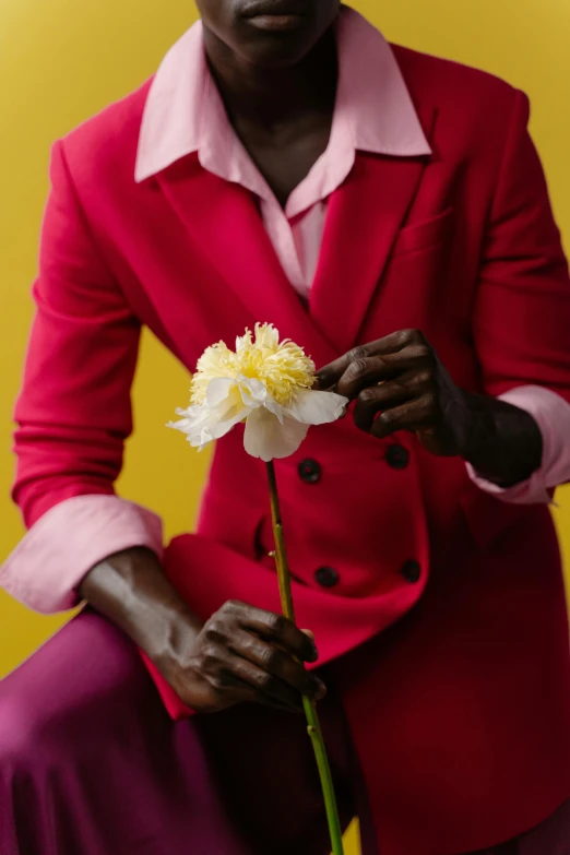 a man sitting on a chair holding a flower, by Alison Geissler, trending on pexels, raspberry banana color, adut akech, red suit, pink and yellow