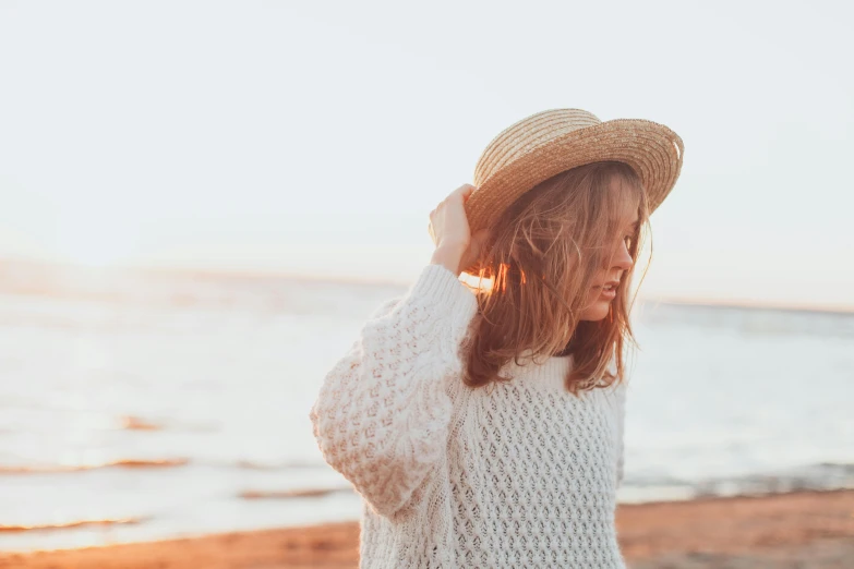 a woman standing on top of a beach next to the ocean, pexels contest winner, knitted hat, wearing a white sweater, soft vintage glow, background image