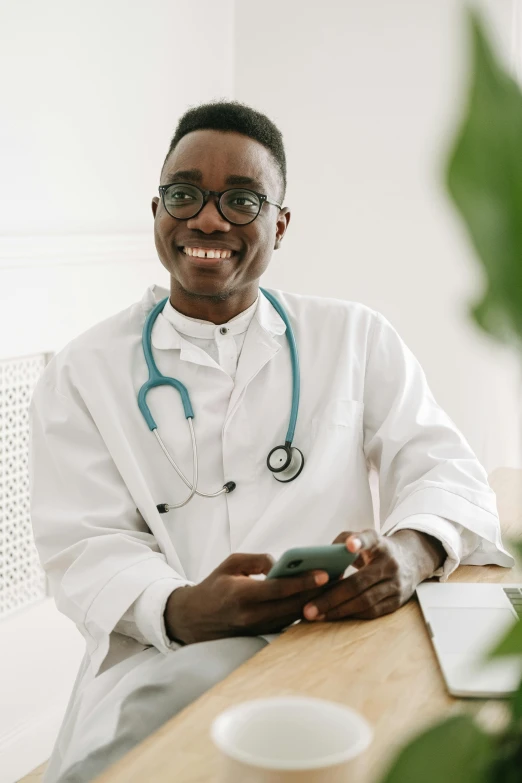 a close up of a person sitting at a table with a cell phone, lab coat and tee shirt, stethoscope, looking happy, lgbtq