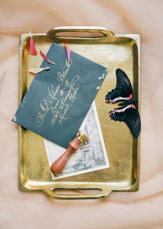 a black butterfly sitting on top of a gold tray, inspired by Eden Box, featured on instagram, private press, with some hand written letters, custom brush, a wooden, a brightly colored