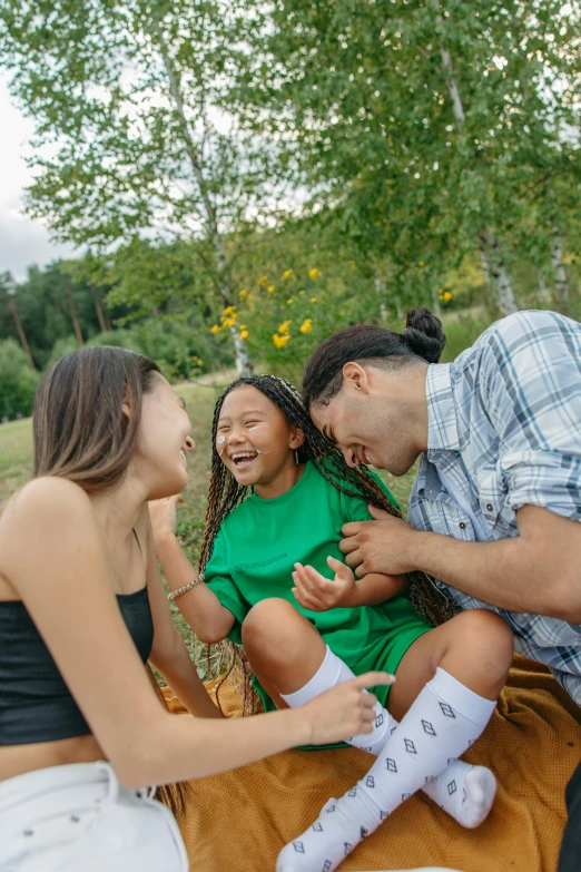 a couple of people that are sitting on a blanket, with a kid, laughing, in a open green field, gen z