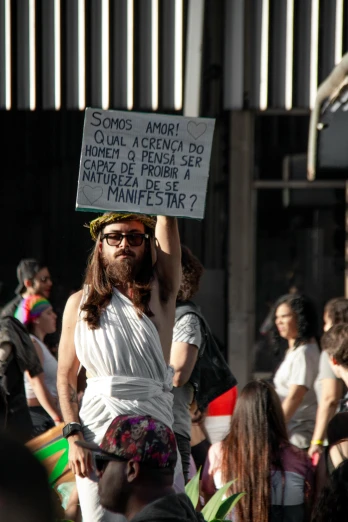 a man with a sign in the middle of a crowd, by Ramón Silva, reddit, renaissance, wearing a toga and sandals, chilean, drag, in the sun