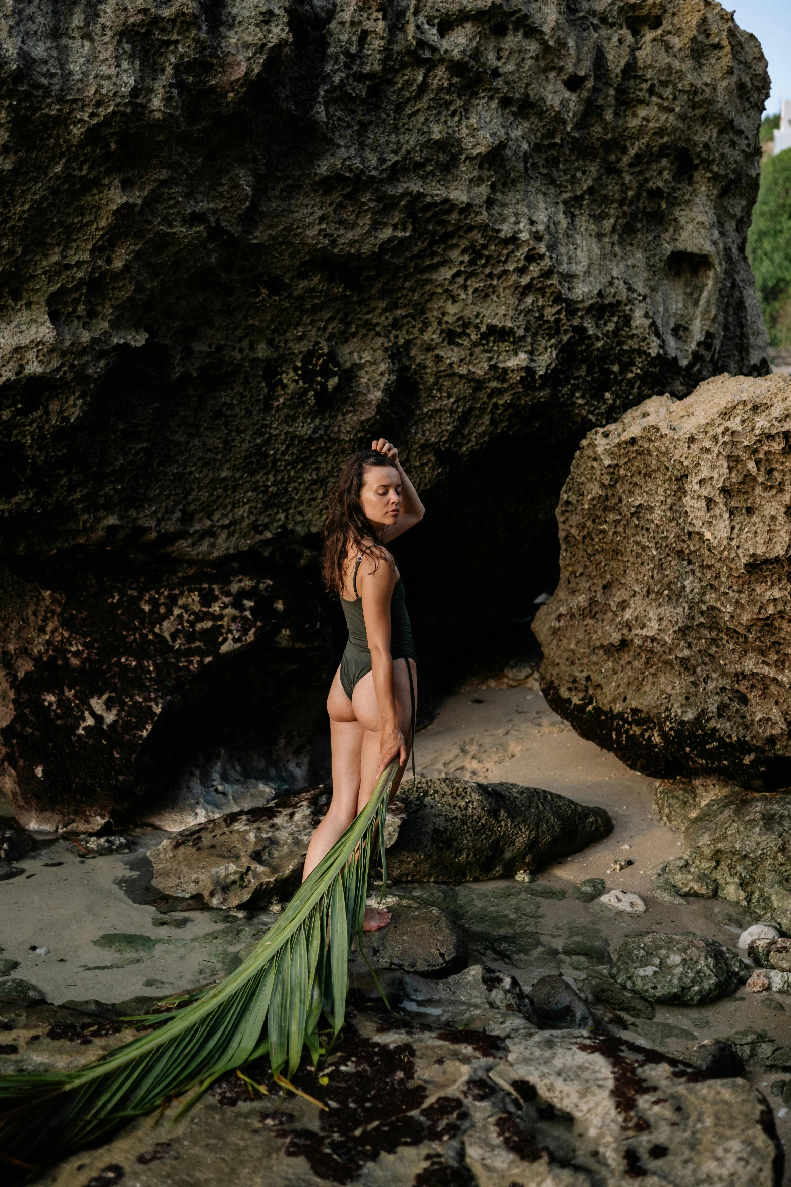 a woman standing next to a large rock on a beach, an album cover, unsplash, sumatraism, green swimsuit, vines, asian women, crouching