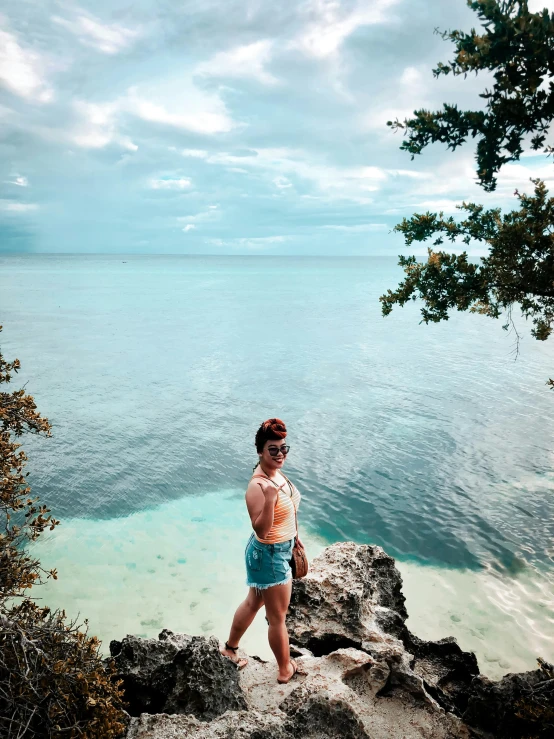 a woman standing on top of a rock next to the ocean, carribean turquoise water, profile image, beach trees in the background, instagram photo