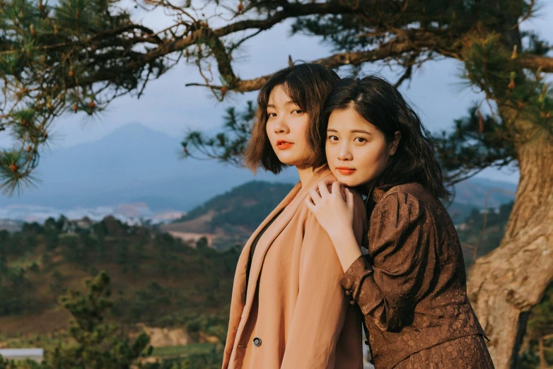 two women standing next to each other near a tree, an album cover, inspired by Itō Shinsui, trending on pexels, sumatraism, hills in the background, portrait shot 8 k, vietnamese woman, brunettes