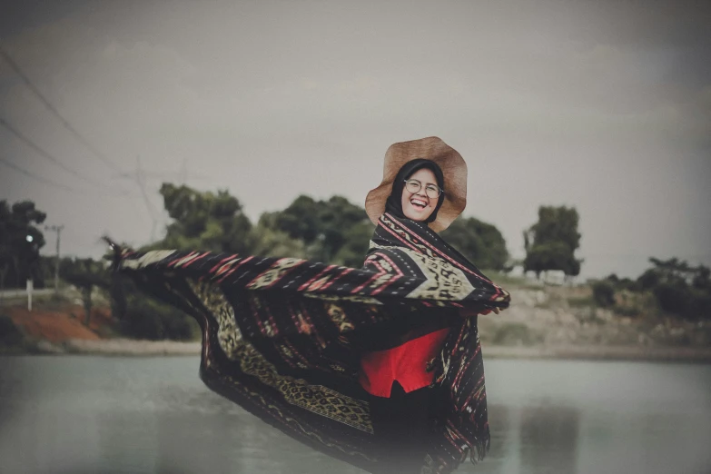 a woman standing next to a body of water, by Ismail Acar, pexels contest winner, hurufiyya, covered with blanket, smiling and dancing, retro effect, iraq nadar