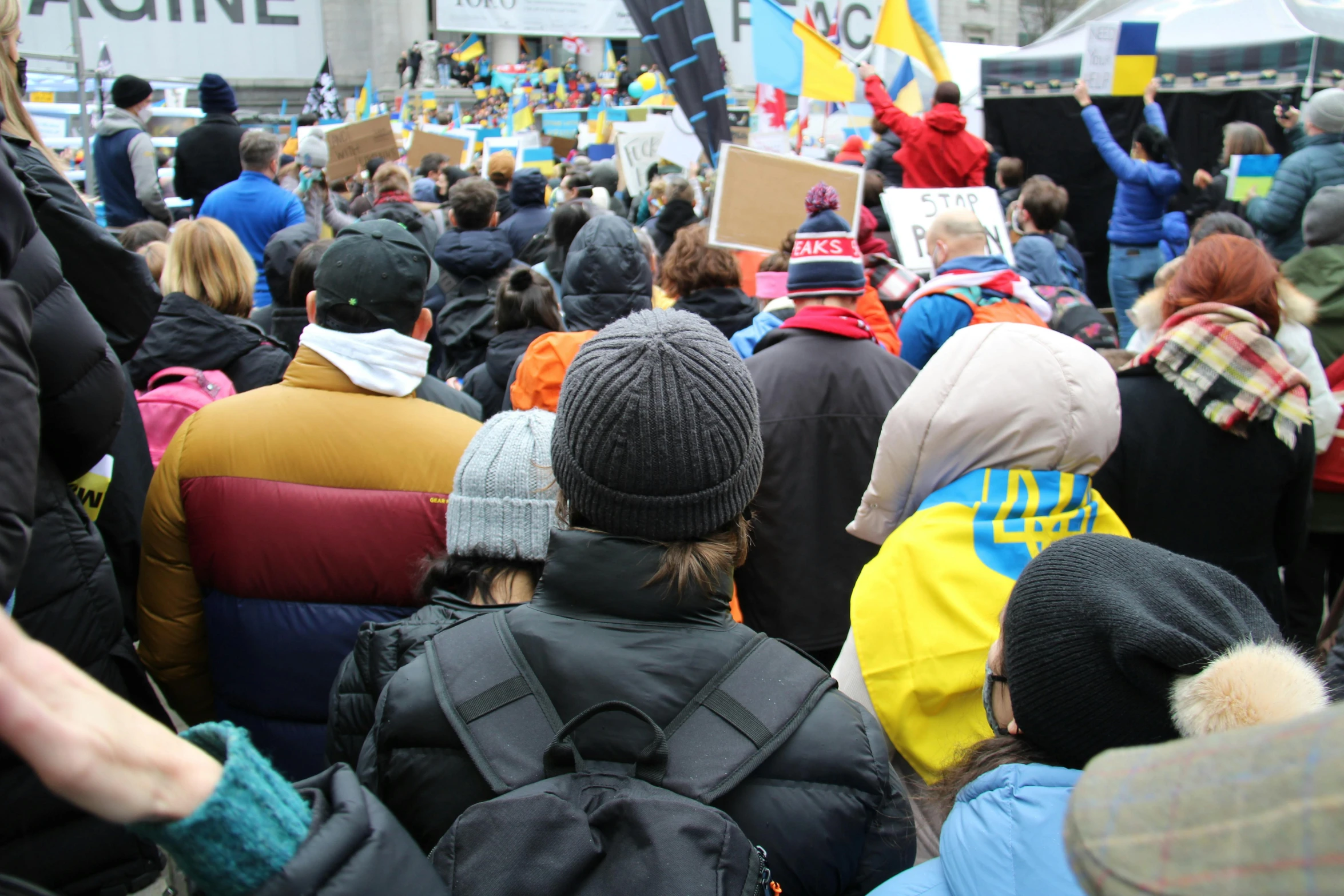 a group of people that are standing in the street, yellow and blue and cyan, russian opposition rally, seen from behind, square