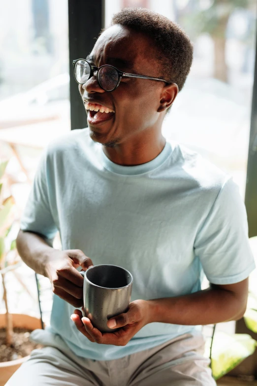a man sitting in front of a window holding a cup, pexels contest winner, happening, earing a shirt laughing, black man, two cups of coffee, wearing black glasses