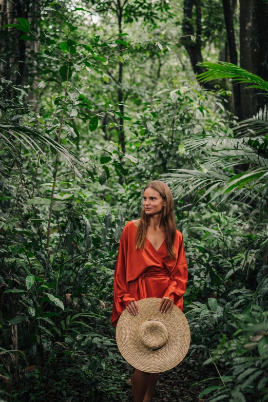 a woman standing in the middle of a forest, by Emma Andijewska, sumatraism, orange jumpsuit, tropical setting, wearing wide sunhat, wearing a luxurious silk robe