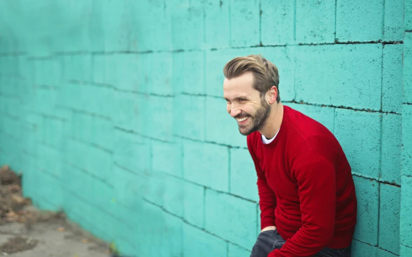 a man leaning against a blue brick wall, happening, red sweater and gray pants, smiling playfully, with teal clothes, short dark blond beard