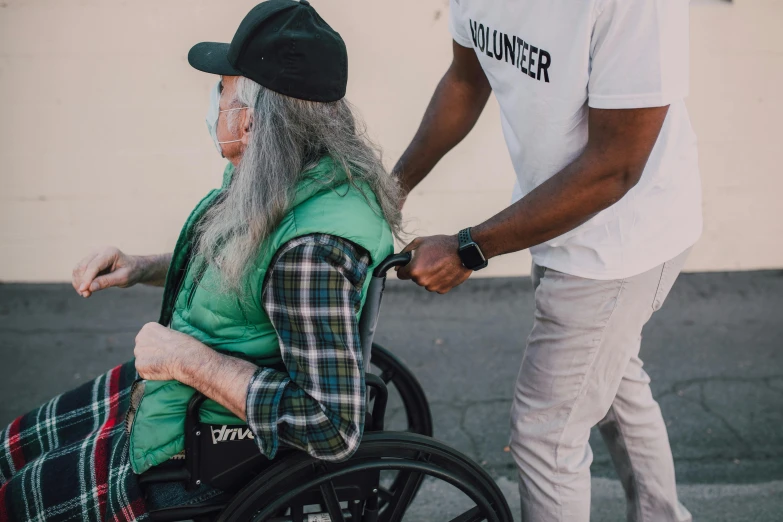 a man standing next to a woman in a wheelchair, by Matija Jama, pexels contest winner, renaissance, wearing a baggy, profile image, wounded, acronym