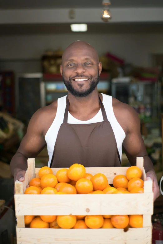 a man holding a crate full of oranges, a portrait, pexels, ( ( dark skin ) ), wearing an apron, convenience store, muscular bald man