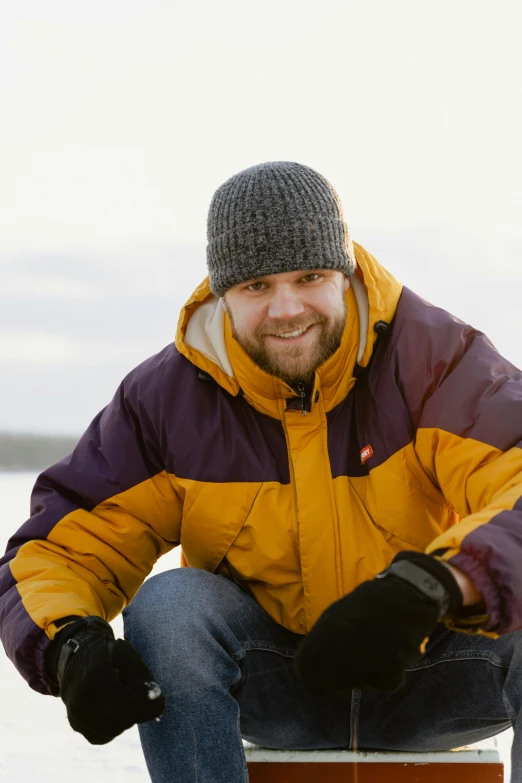 a man sitting on a box in the snow, wearing a purple cap, nina tryggvadottir, near a lake, yellow