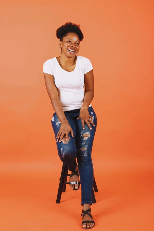 a woman sitting on top of a wooden chair, by Stokely Webster, happening, wearing jeans, in front of an orange background, laughing, white backdrop