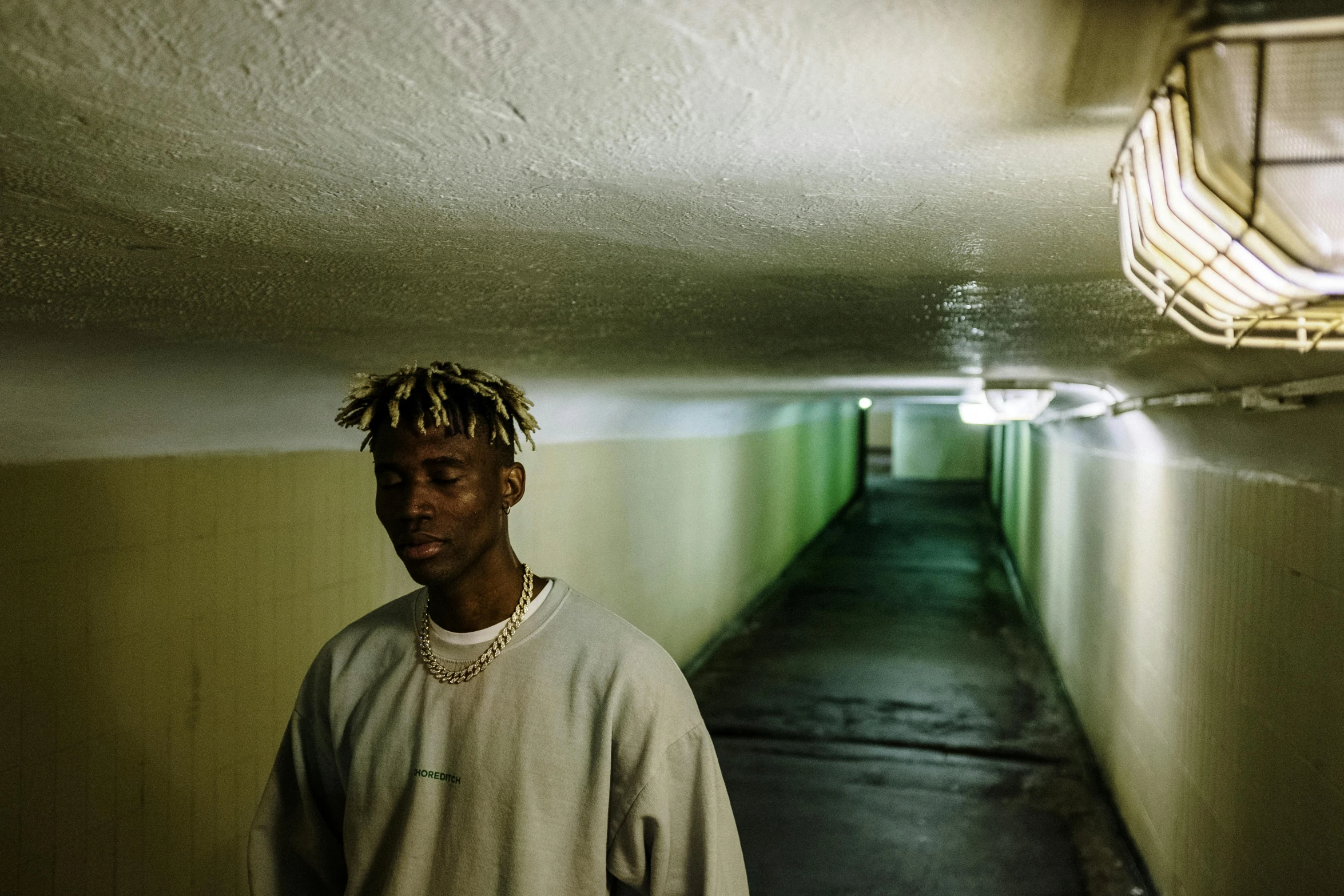 a man with dreadlocks standing in a hallway, inspired by Elsa Bleda, pexels contest winner, les nabis, tyler the creator, wearing a gold chain, in an underground parking garage, wearing adidas clothing