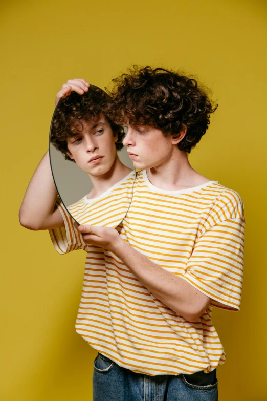 a couple of young men standing next to each other, an album cover, trending on pexels, magic realism, wavy hair yellow theme, looking into a mirror, sophia lillis, genderless