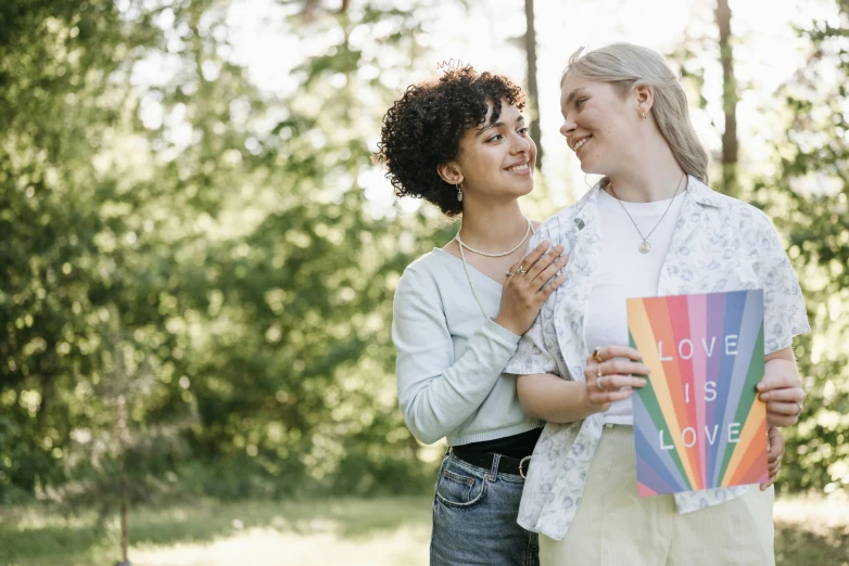 a couple of women standing next to each other, pexels, happening, pride month, holding a book, love os begin of all, outdoor photo