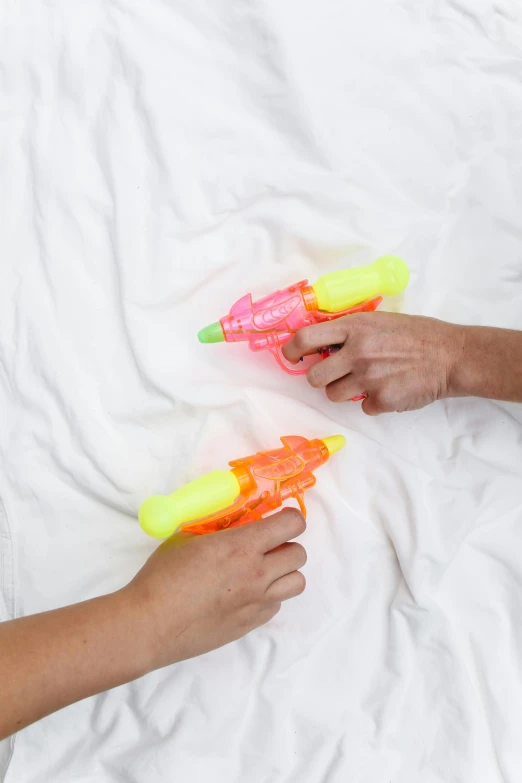 two people are playing with water guns on a bed, zoomed out shot, laying down with wrists together, neons, product shot