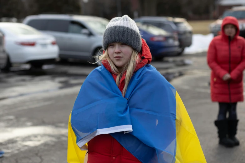 a woman wrapped in a blanket walking down a street, ukrainian flag on the left side, erin moriarty, wearing a red backwards cap, 2019 trending photo