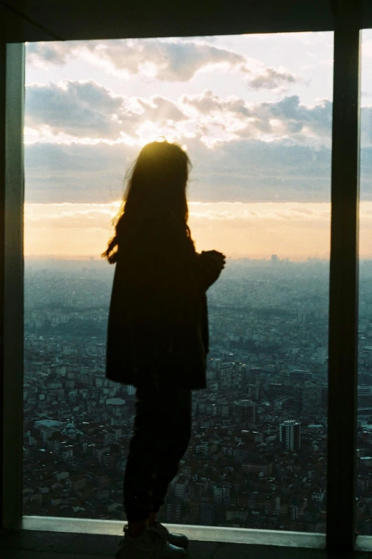 a person standing in front of a window looking out at a city, by Eizan Kikukawa, happening, silhouette over sunset, panoramic view of girl, taken in the late 2010s, sky - high view