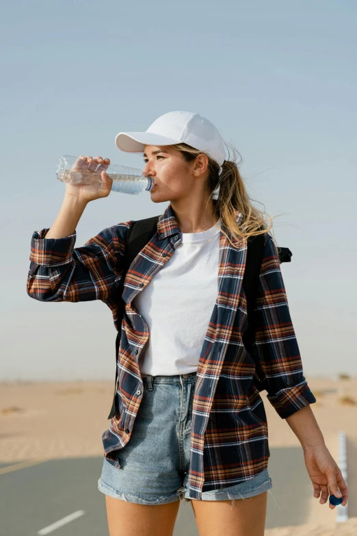 a woman standing on the side of a road drinking water, wearing a flannel shirt, standing in desert, detailed product image, wearing baseball cap