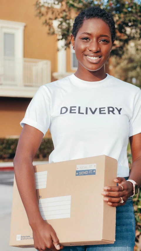 a woman holding a box in front of a building, wearing a t-shirt, dark skinned, ecommerce photograph, digital image