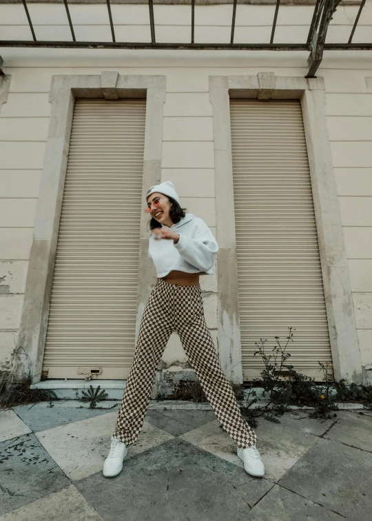 a woman standing in front of a building talking on a cell phone, an album cover, trending on pexels, wearing pants, checkered motiffs, wearing a white sweater, playful pose