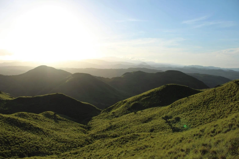 a person flying a kite on top of a lush green hillside, sumatraism, sun setting, sparse mountains on the horizon, shades of green, instagram post