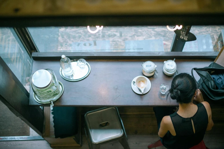 a woman sitting at a table in front of a window, trending on unsplash, bird's eye view, mysterious coffee shop girl, secret tea society, from back