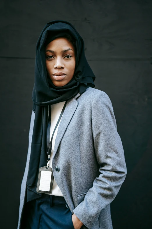 a woman standing in front of a black wall, by Alasdair Grant Taylor, hurufiyya, grey jacket, riyahd cassiem, black scarf, office clothes