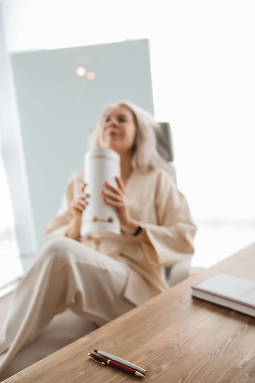 a woman sitting in a chair with a cup of coffee, by Matija Jama, pexels contest winner, long wavy white hair, carrying a bottle of perfume, wearing white pajamas, sitting in office