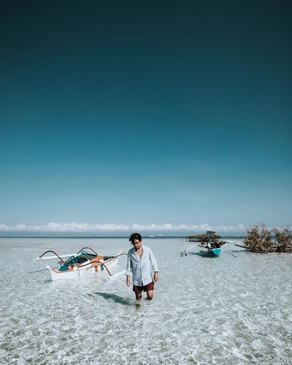 a man standing on top of a sandy beach, by Adam Dario Keel, pexels contest winner, sumatraism, wearing white cloths, boats in the water, crystal clear blue water, carrying a tray