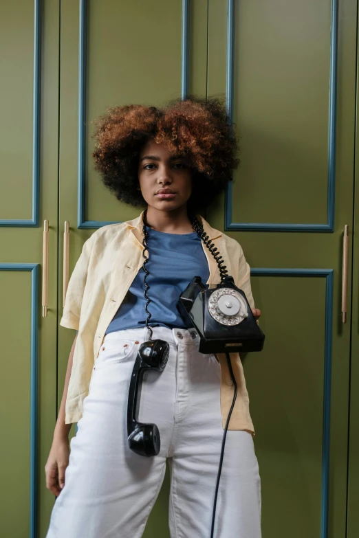 a woman standing in front of a green wall holding a phone, a portrait, inspired by Nan Goldin, trending on pexels, afrofuturism, 1960s style, dials, loose - fitting blouses, black teenage girl