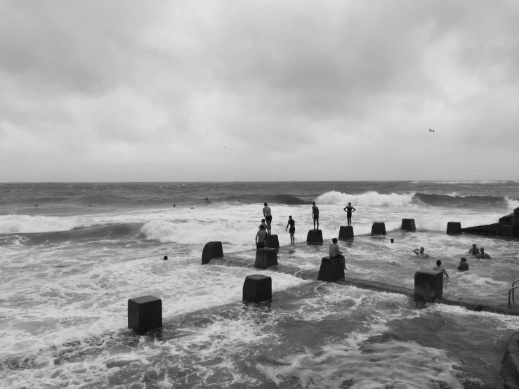 a group of people standing on top of a beach next to the ocean, a black and white photo, by Colijn de Coter, unsplash, surrealism, violent stormy waters, wooden platforms, people swimming, the sea seen behind the city