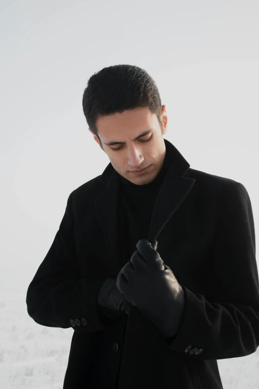 a man in a black coat standing in the snow, an album cover, inspired by Nadim Karam, black leather gloves, profile shot, clean shaven, official store photo