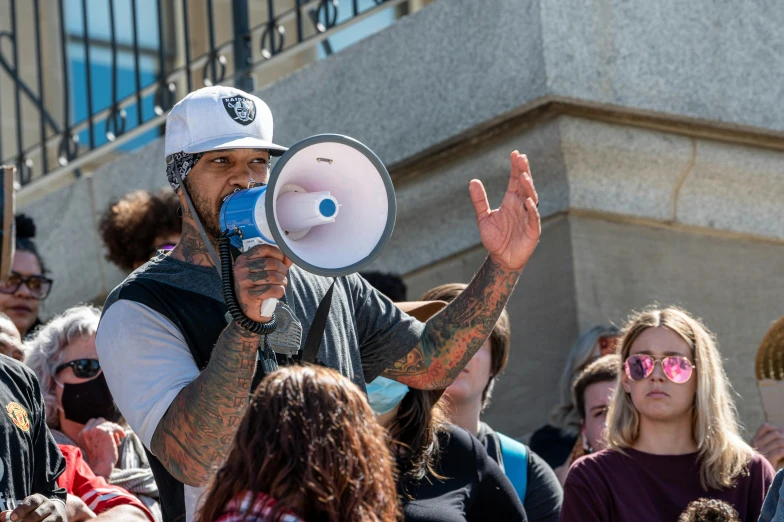 a man holding a megaphone in front of a crowd, a photo, square, brandon, 2022 photograph, gray