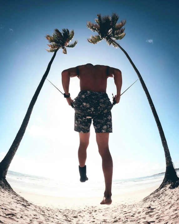 a man standing on a beach next to two palm trees, by Jessie Algie, pexels contest winner, happening, athletic shorts, mesomorph, trapped on a hedonic treadmill, arched back