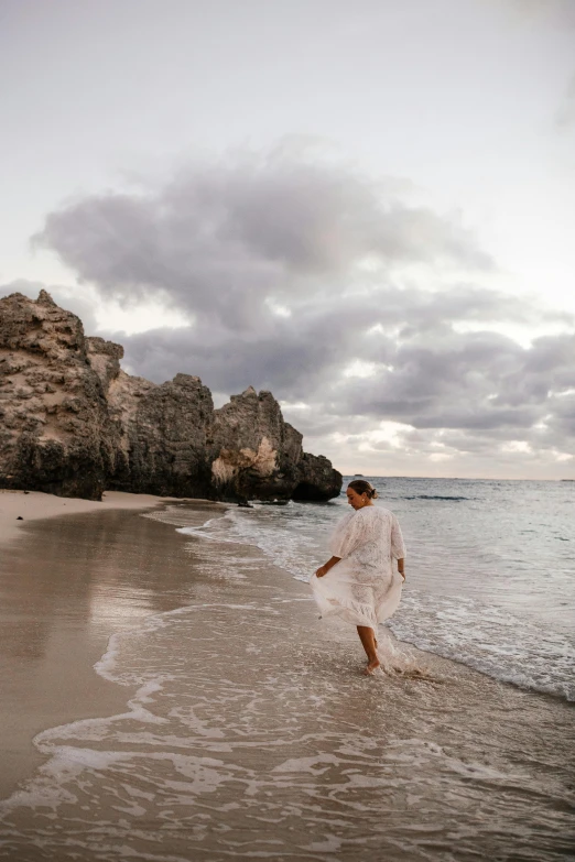 a woman walking on a beach next to the ocean, rocky cliff, floating robes, carribean white sand, grey