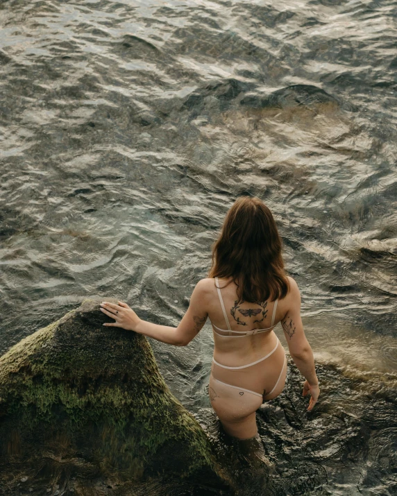 a woman in a bikini sitting on a rock in the water, inspired by Elsa Bleda, unsplash contest winner, renaissance, looking from behind, queer woman, currents, her skin is light brown