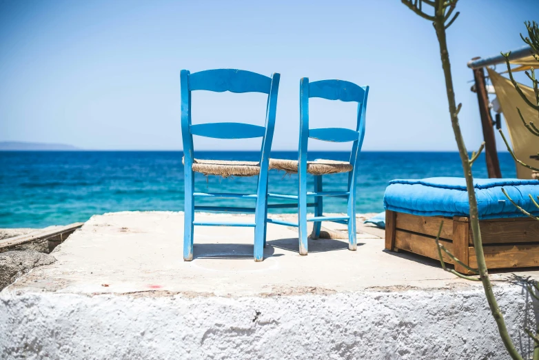 two blue chairs sitting on top of a rock next to the ocean, a still life, pexels contest winner, romanticism, greek style, summer street near a beach, un restaurant avec une terrasse, uncropped