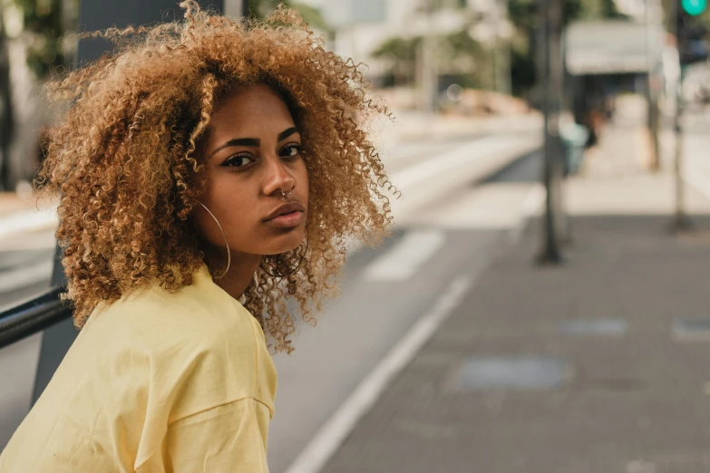 a woman with curly hair on a city street, trending on pexels, realism, yellow and black, young women, short blonde afro, distant expression