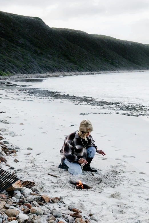 a person kneeling on a beach next to a body of water, a picture, with some sausages on the fire, cape, coastline, chilly