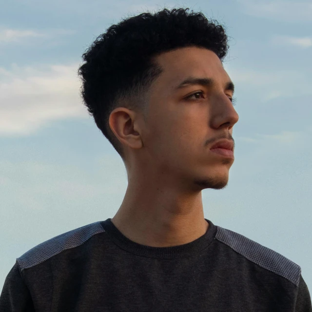 a young man standing in front of a blue sky, an album cover, inspired by Carlos Berlanga, pexels contest winner, realism, mixed race, backlit face, raden saleh, looking serious