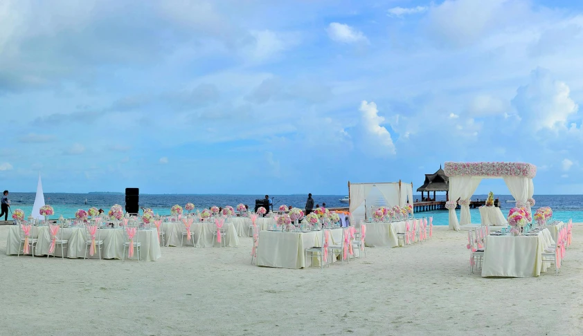 a group of people standing on top of a sandy beach, flower decorations, white and pink cloth, tables and chairs, square