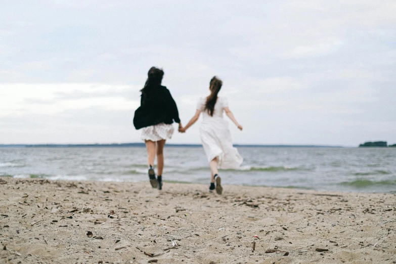 a couple of women walking on top of a sandy beach, pexels contest winner, romanticism, girl is running, wearing a white dress, good friends, slight overcast