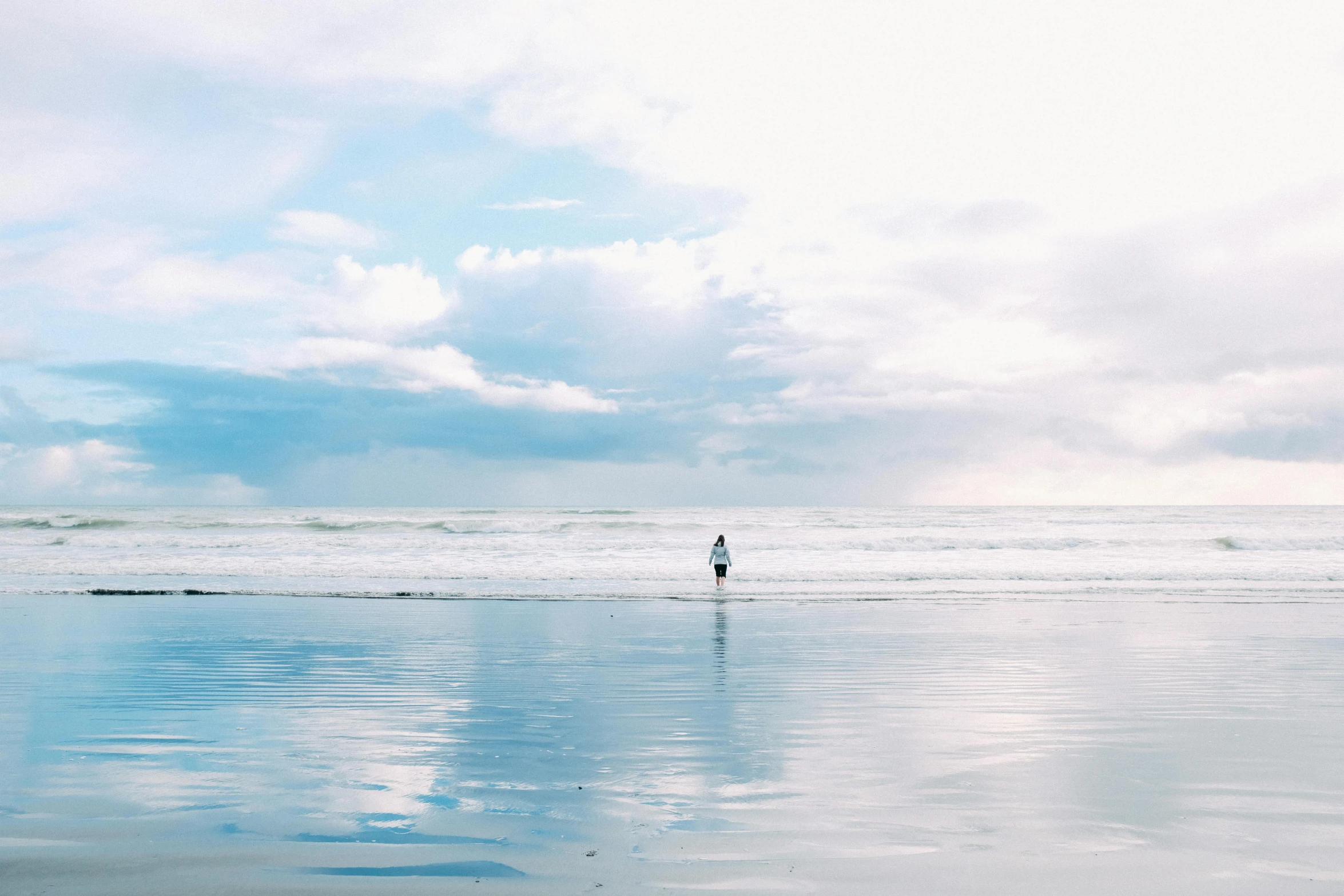 a person standing on top of a beach next to the ocean, unsplash contest winner, minimalism, glossy reflections, new zealand, background image, cloudy skies