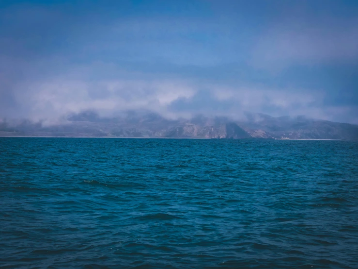 a large body of water with a mountain in the background, an album cover, unsplash, blue fog, hollister ranch, offshore winds, ignant