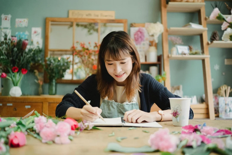 a woman sitting at a table writing on a piece of paper, a drawing, inspired by Masamitsu Ōta, pexels contest winner, flower shop scene, avatar image, portrait photo, paper quilling