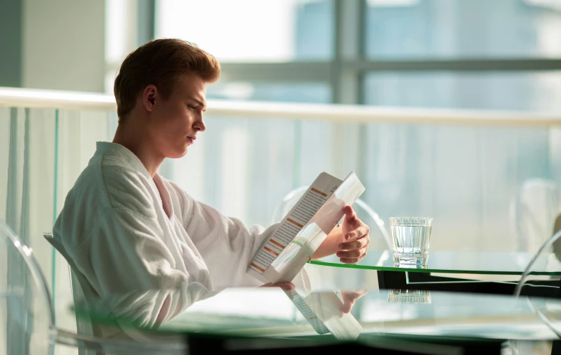 a woman sitting at a table reading a book, hyperrealism, wearing a white hospital gown, androgynous male, hotel room, reading engineering book